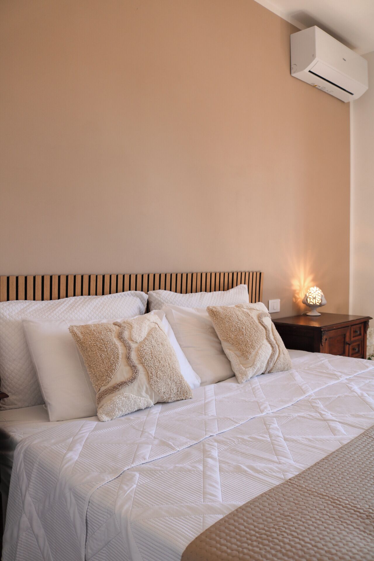 Un letto rifatto con cura, lenzuola bianche e cuscini soffici, una testiera in legno, un comodino con una lampada e un condizionatore d'aria a parete.