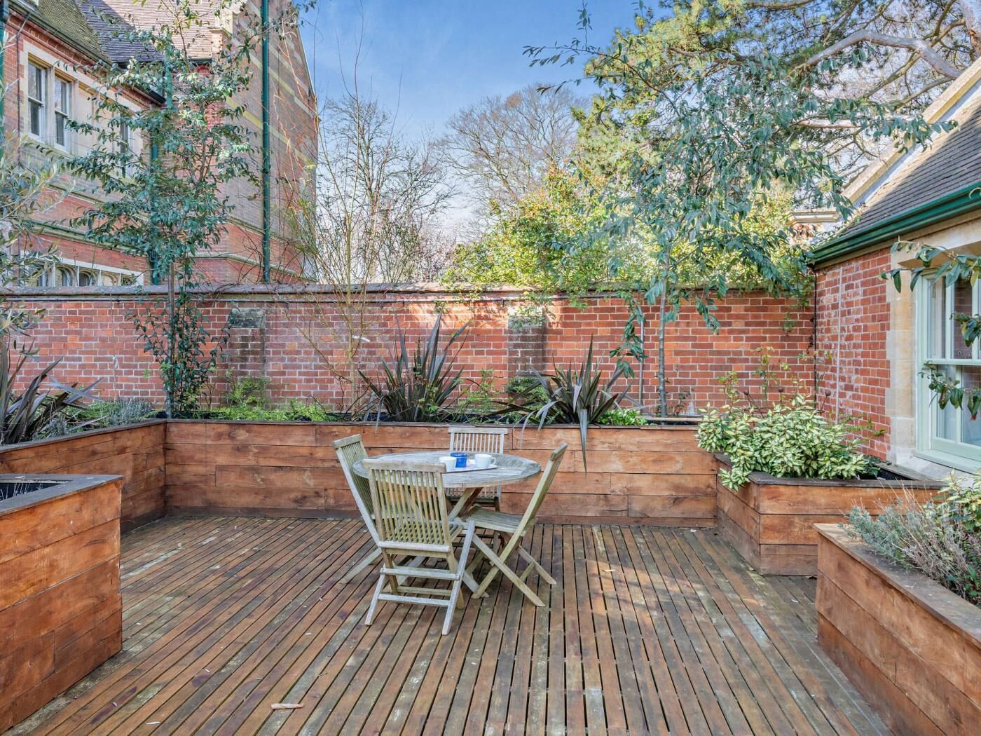 A wooden deck with a table and chairs, surrounded by brick walls and various plants.