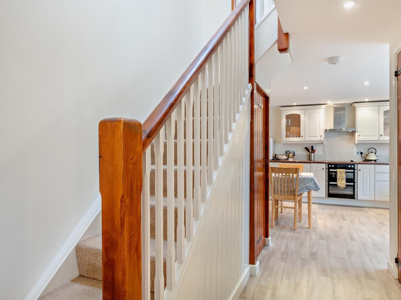 A staircase with wooden handrails leading to a kitchen area with white cabinets and a dining table.