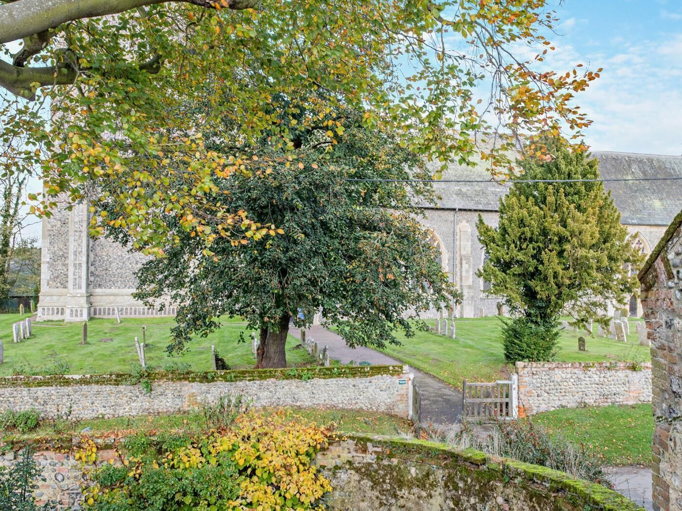 A historic building with a large tree in the foreground, a stone wall, and a grassy area with headstones.
