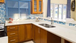 A kitchen with wooden cabinets, a white countertop, a stainless steel sink, and a window with blinds. - Coolshade - Richmond Estate  (St. Ann Parish)
