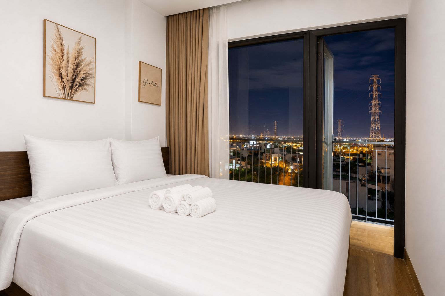 A hotel room with a bed, pillows, a view of the city at night, and a framed picture on the wall.