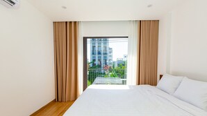 Habitación de hotel con una cama grande, un balcón con vistas a edificios y follaje, y una ventana con cortinas.