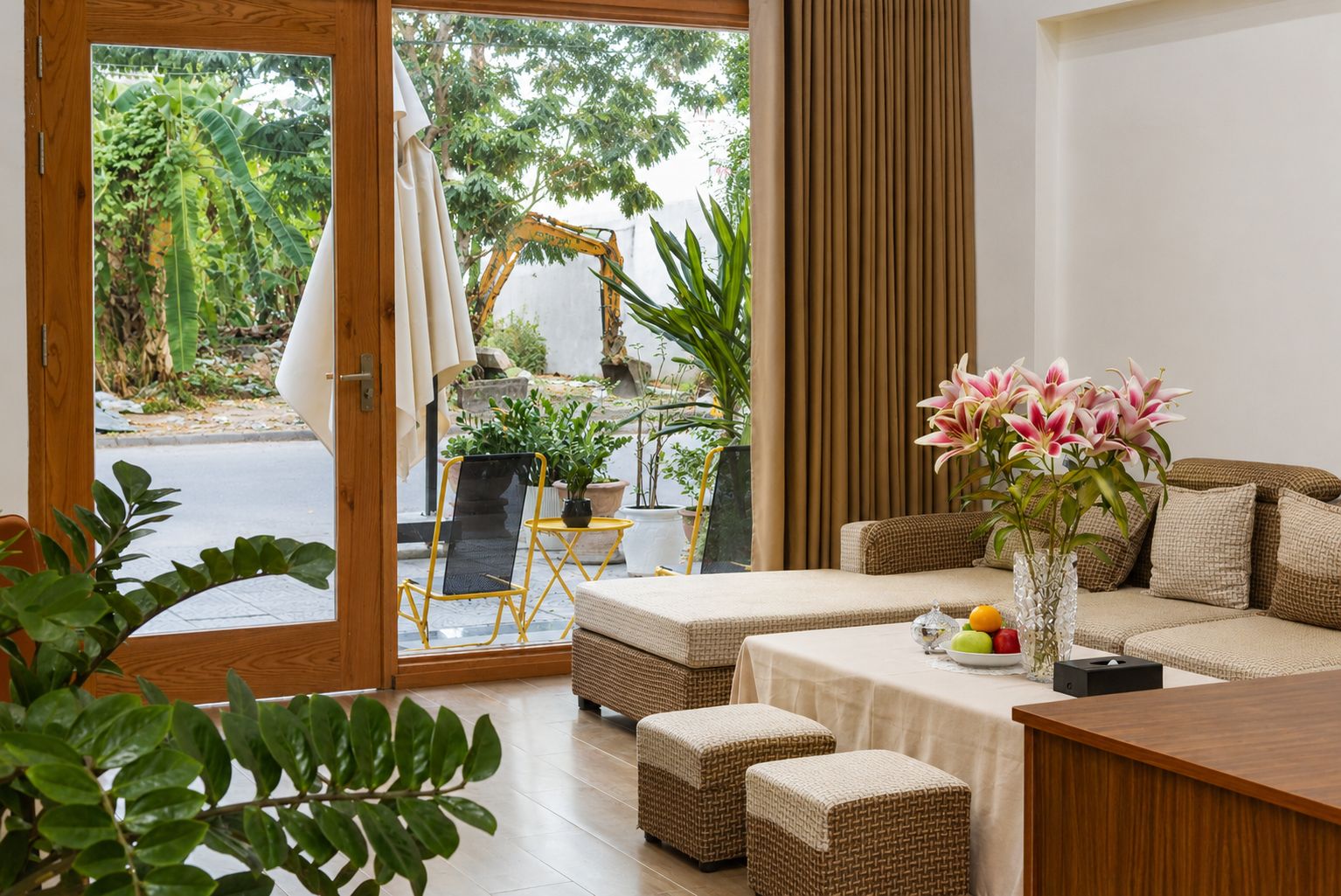 A room with a sofa, ottomans, a table with fruit and a vase, a chair, and a view of greenery and an excavator outside.