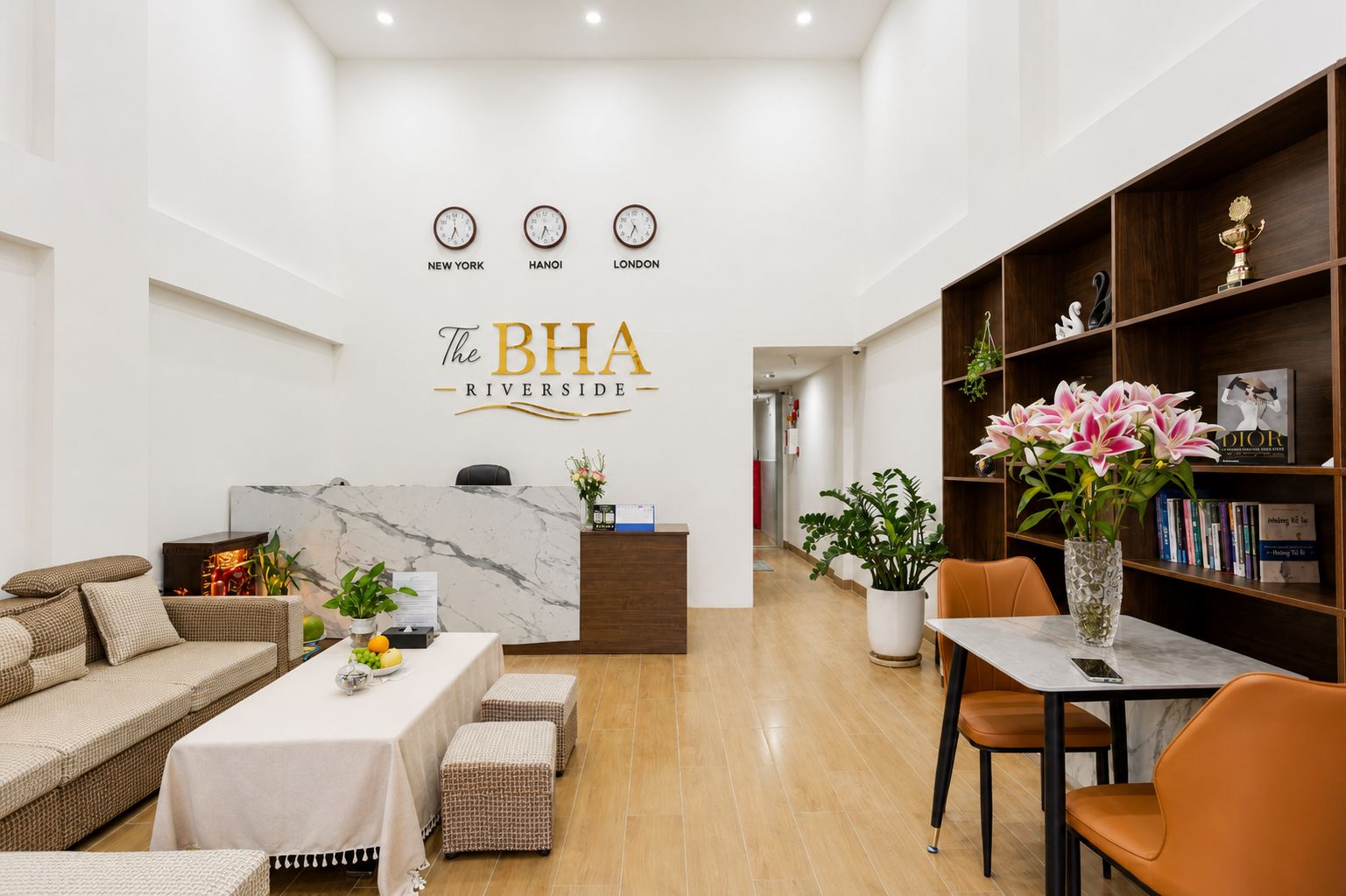 A hotel reception area with a marble reception desk, a sofa, a dining table with chairs, and a bookshelf with books and flowers.