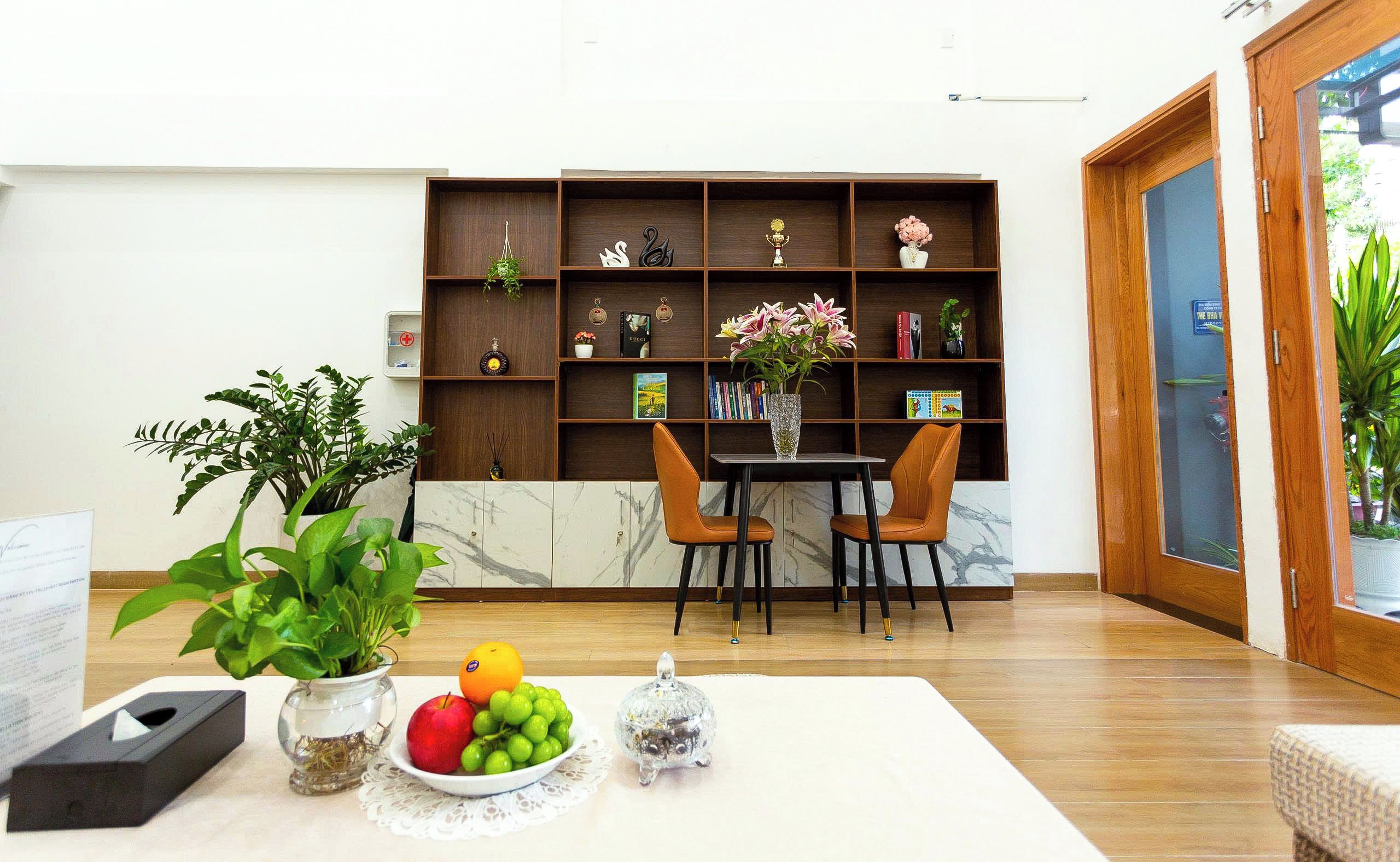 A room with a wooden bookshelf, a marble-topped table with fruit and a vase, two orange chairs, and a potted plant.