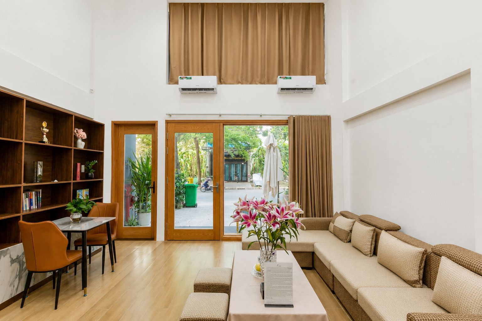 A spacious living area with a sofa, a dining table, and a bookshelf. There are two sliding glass doors leading to an outdoor area.