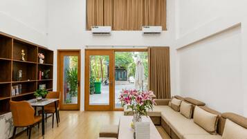 A spacious living area with a sofa, a dining table, and a bookshelf. There are two sliding glass doors leading to an outdoor area.