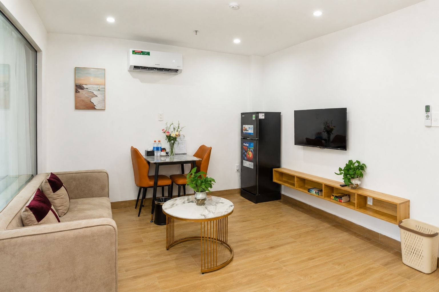 A modern living room with a sofa, dining table, chairs, a TV, a refrigerator, and a small plant on a shelf.