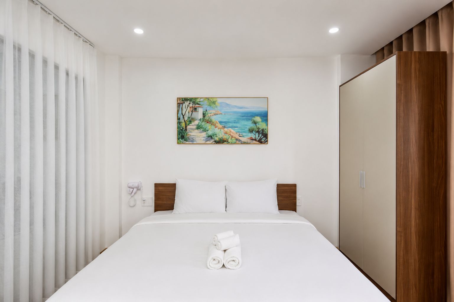 A hotel room with a bed, a painting of a beach scene, a wooden wardrobe, and a hairdryer on the wall.