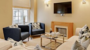 Ein Wohnzimmer mit Kamin, zwei Sesseln, einer Couch und einem Couchtisch. Ein Fernseher ist an der Wand befestigt.