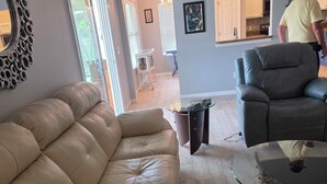Ein Wohnzimmer mit Ledersofa, Glastisch und dunklem Sessel. Im Hintergrund ist ein Essbereich erkennbar.