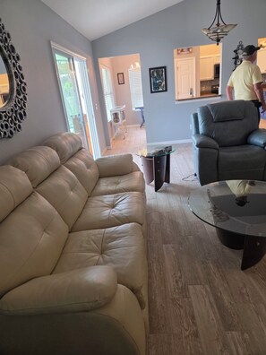 Ein Wohnzimmer mit Ledersofa, Glastisch und dunklem Sessel. Im Hintergrund ist ein Essbereich erkennbar.