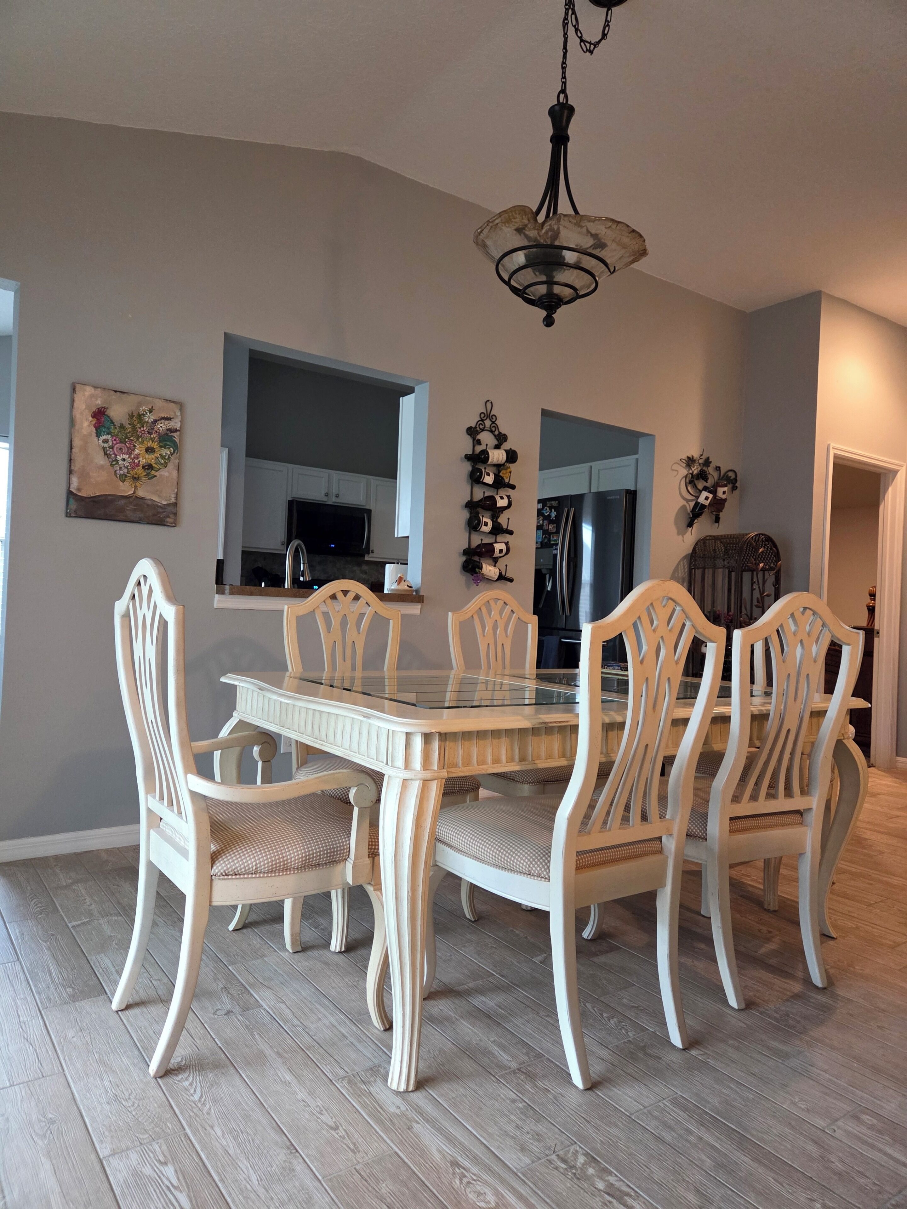 A dining area with a round glass table surrounded by six chairs, a chandelier, and a kitchen in the background.