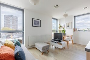 Ein modernes Wohnzimmer mit einem großen Fenster, einem Flachbildfernseher, einem kleinen Tisch und einem Sofa mit bunten Kissen.
