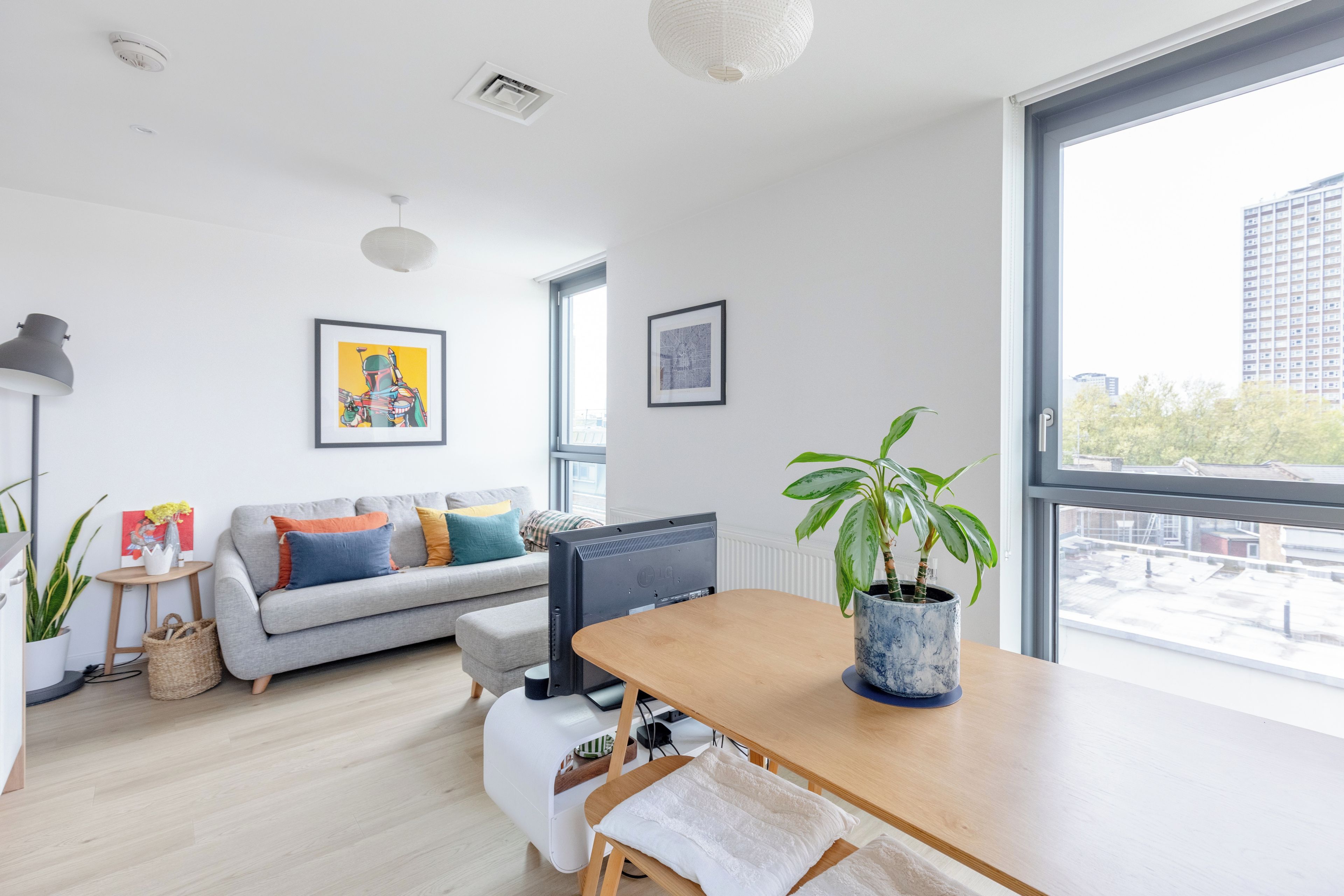 Ein modernes Wohnzimmer mit einem grauen Sofa, einem Holzesstisch, einem großen Fenster und einer Zimmerpflanze.