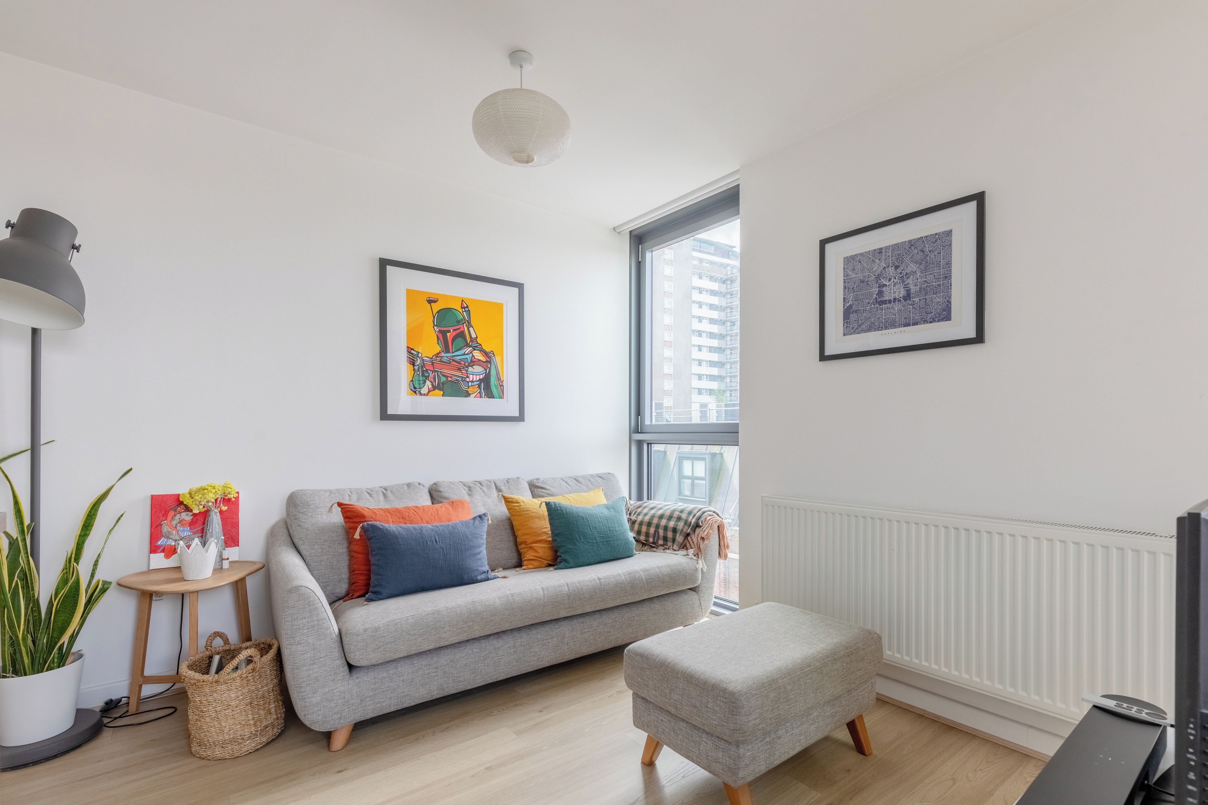 Ein modernes Wohnzimmer mit einem grauen Sofa, bunten Kissen, einem kleinen Beistelltisch mit einer Vase, einem großen Fenster und einem gerahmten Stadtbild.