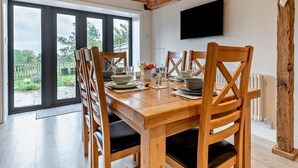 Een eetruimte met een houten tafel en stoelen, een aan de muur bevestigde televisie en grote ramen met uitzicht op groen.