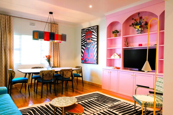 A modern living room with a dining area, a flat-screen TV, and a pink shelving unit. - Designer Family Home with Garden & Workspace – Walking Distance to the Beach (Cape Town)