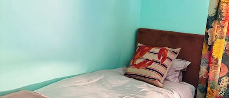 Uma cama de solteiro com cabeceira vermelha, almofada estampada e manta vermelha. Um banquinho amarelo está posicionado aos pés da cama.