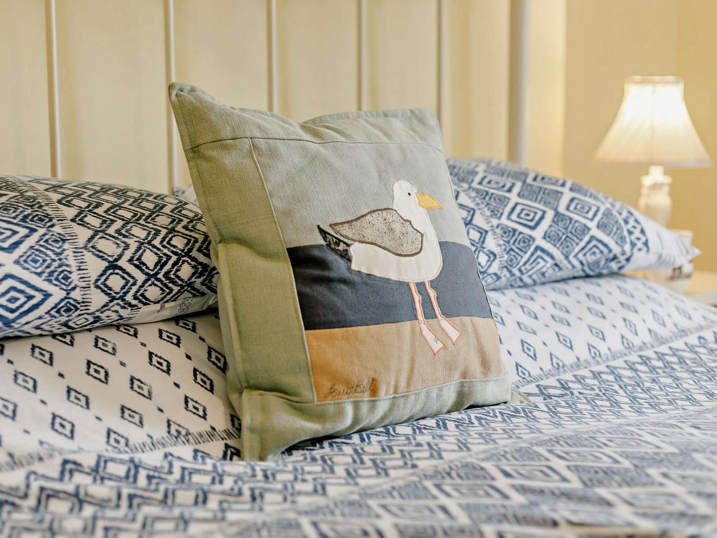 En seng med mønstret sengetøj, en pude med et design af en måge og en lampe ved siden af.