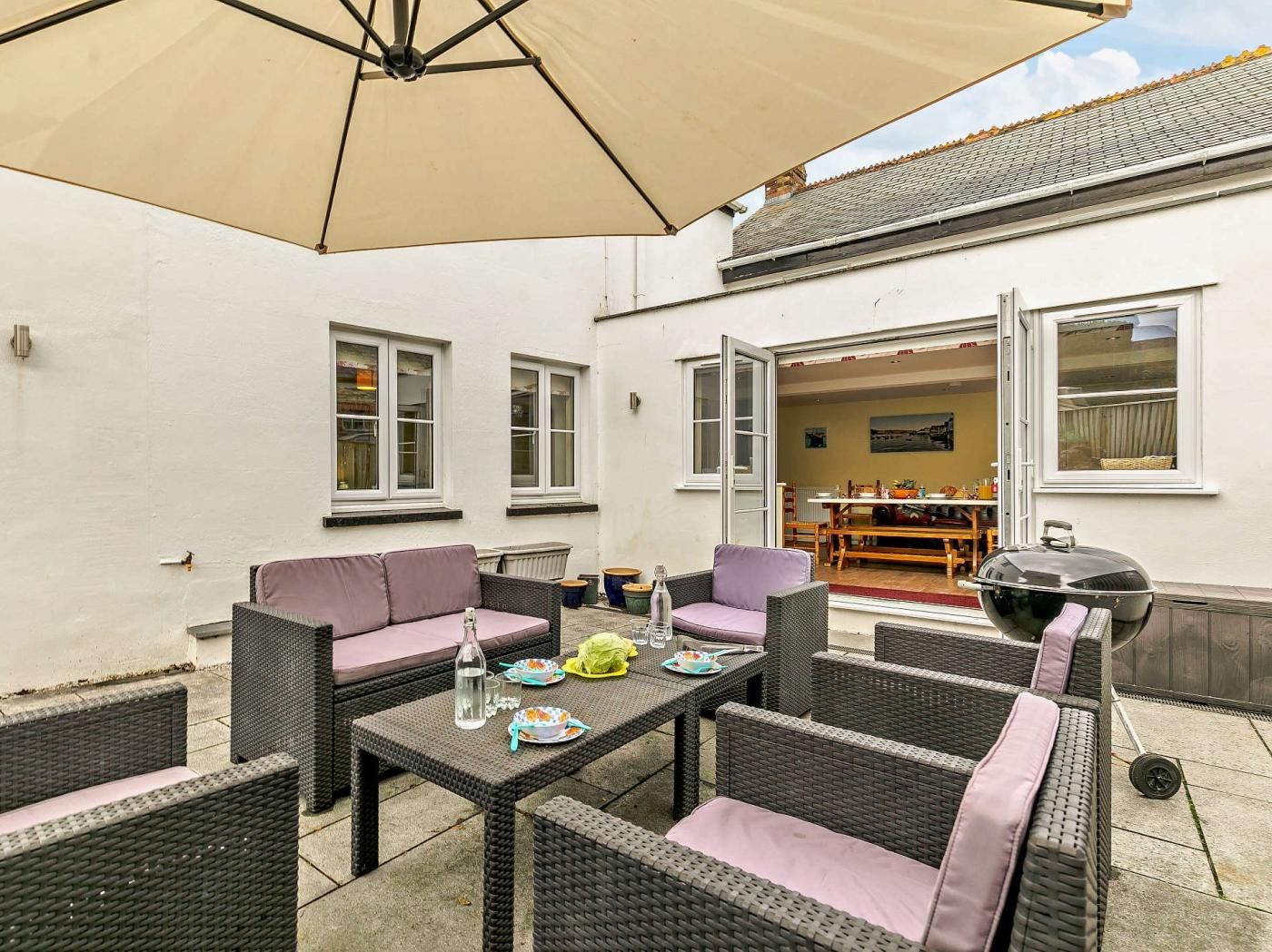 Een terras buiten met rieten meubels, een tafel gedekt met borden en glazen, en een barbecue.