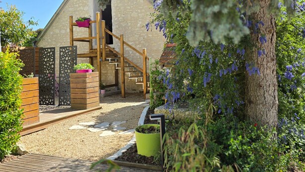 Una escalera de madera lleva a un edificio de piedra con techo inclinado, rodeado por un jardín con terraza de madera y plantas en macetas.