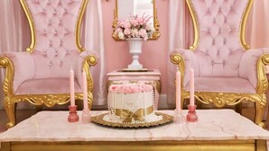 Ein luxuriöser Raum mit einem Marmortisch, zwei in Rosa gepolsterten Sessel, einer dekorativen Torte und einer Blumenvase.