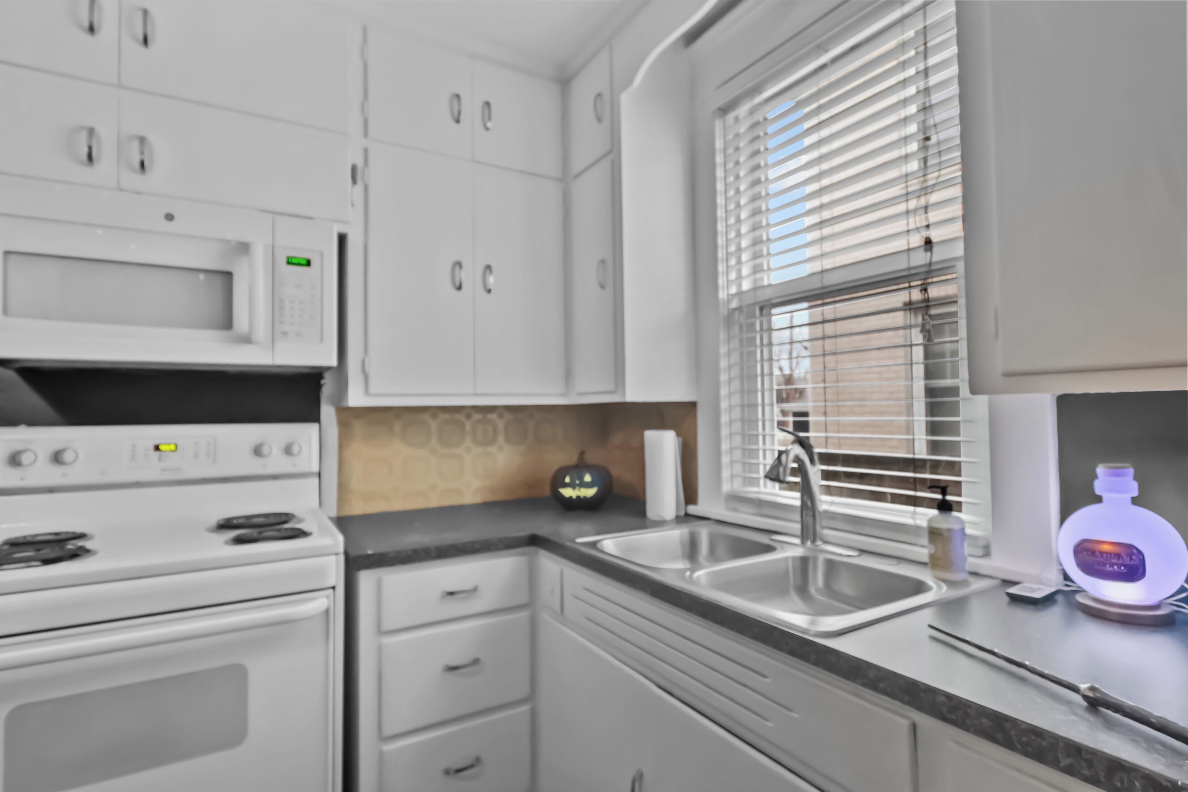 Une cuisine moderne avec des meubles blancs, un four à micro-ondes, une cuisinière, un évier et une fenêtre avec des stores.