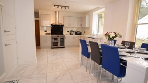A modern kitchen with white cabinetry, a central island, and a dining area with blue chairs. - 4 Bed in Ilfracombe (oc-66202) (Ilfracombe)