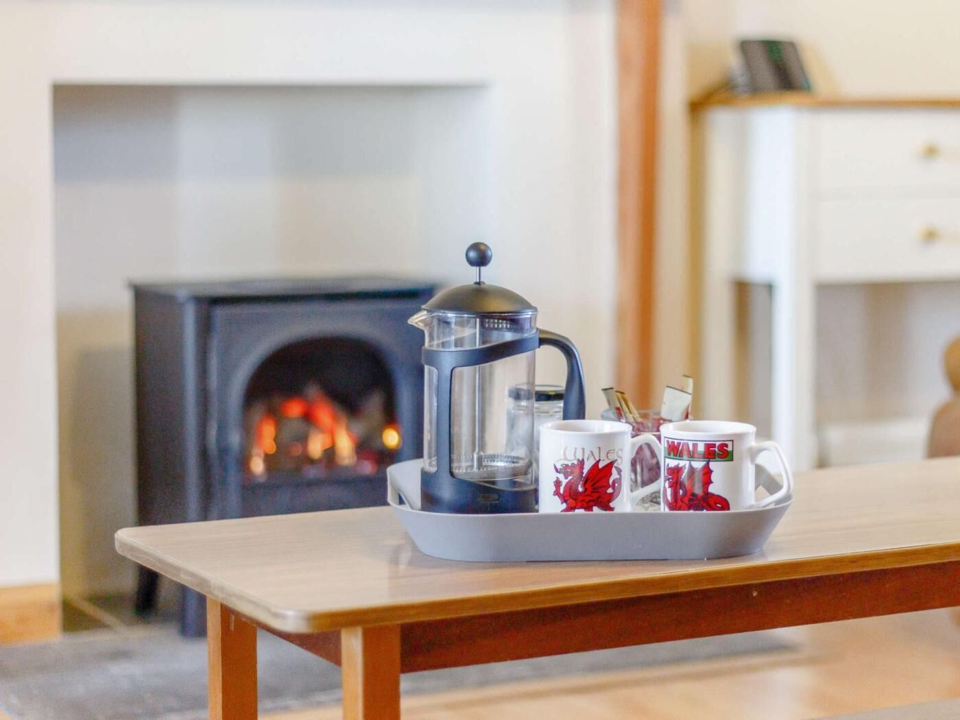 Uma cafeteira sobre uma bandeja com canecas em uma mesa em um quarto com lareira.