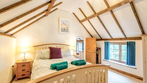 A bedroom with a sloped ceiling, wooden beams, a bed with green towels, a nightstand, a dresser, and a window with curtains. - 2 Bed in South Creake (oc-kt117) (South Creake)