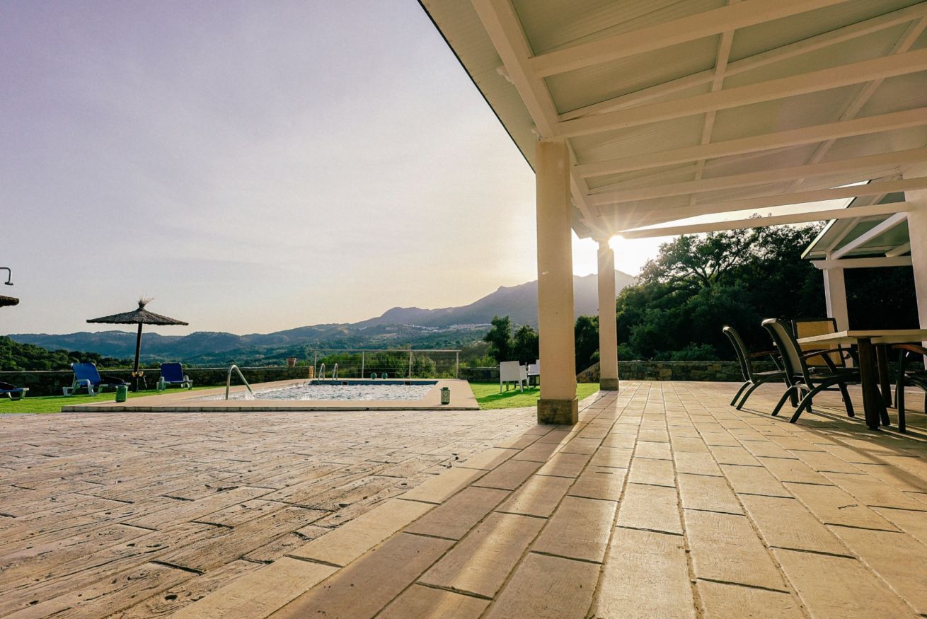 Une terrasse extérieure avec une piscine, des chaises et une table, située sous une zone abritée et offrant une vue sur les montagnes.