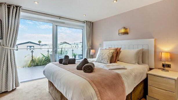 A bedroom with a large bed, a nightstand, a wall-mounted lamp, and a view through a sliding door. - 2 Bed in Bideford (oc-65074) (Northam)