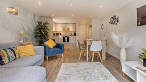 A modern living room with a sofa, a dining table with chairs, a kitchen in the background, and a large plant. - 2 Bed in Bideford (oc-65074) (Northam)