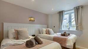 A hotel room with two beds, a bedside table, a lamp, a window with curtains, and a view of the outside. - 2 Bed in Bideford (oc-65074) (Northam)