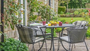 Een ronde metalen tafel met vier stoelen, een stenen gebouw met ramen, en een tuin vol planten en fruit.