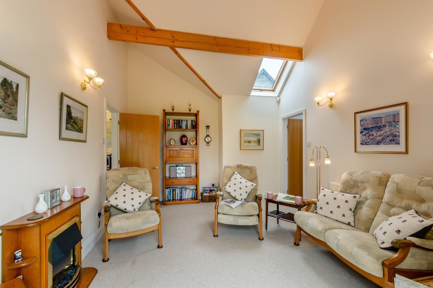 A cozy living room with a fireplace, two armchairs, a sofa, a bookshelf, and a skylight.