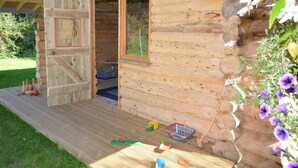 A wooden cabin with a large open door, a window, and a porch area with toys and a basket. - 3 Bed in North Tawton (oc-66043) (Bondleigh)