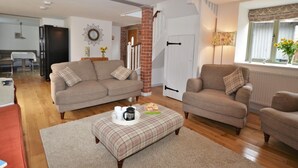 A living room with a sofa, ottoman, and a coffee table with cups and a book. - 3 Bed in North Tawton (oc-66043) (Bondleigh)