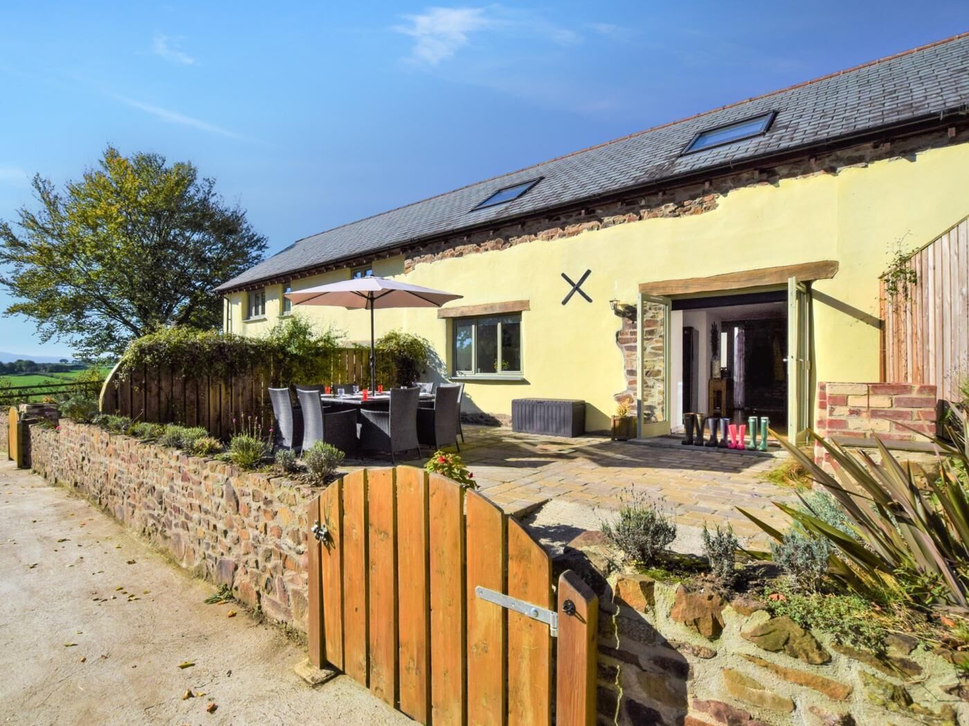 A stone wall, a wooden gate, a patio with outdoor furniture, and a building with a sloped roof.
