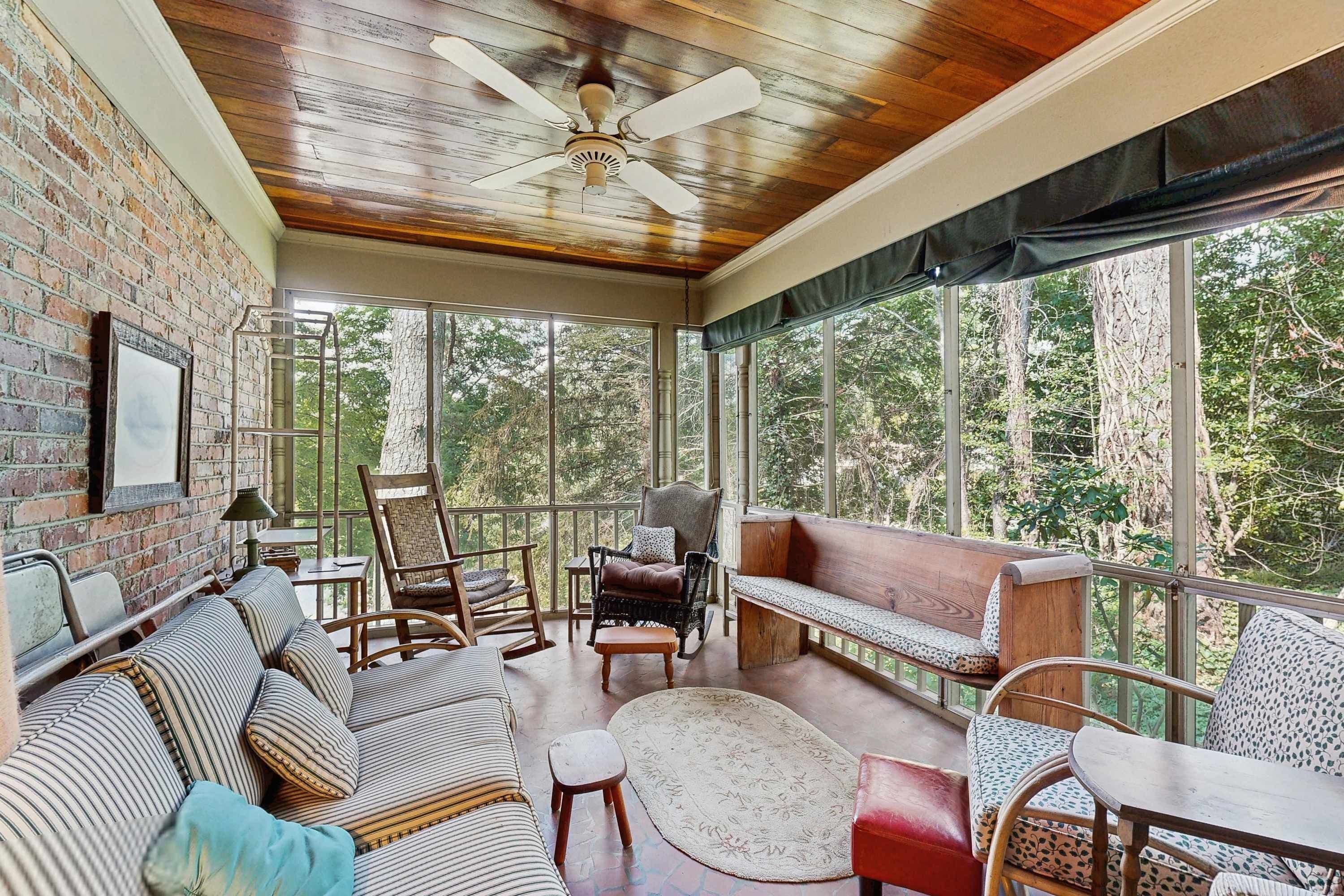 A sunroom with a brick wall, wooden ceiling, and large windows offering a view of trees.
