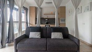 Ein modernes Wohnzimmer mit einem grauen Sofa, einer Küche im Hintergrund und großen Fenstern mit Vorhängen.