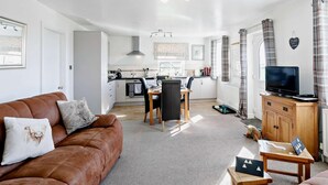 A modern living room with a brown leather sofa, a dining area with a black chair, a kitchen with white cabinets, a wooden cabinet with a TV, and a window with curtains. - 2 Bed in Harrogate (oc-hh018) (Lindley)