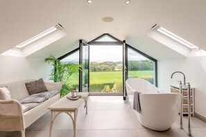 Un intérieur moderne avec une grande verrière, une baignoire blanche et un canapé offrant une vue sur la campagne.