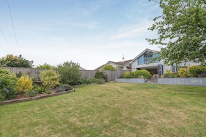 A residential backyard with a lawn, garden beds, and a modern house with a large window and a covered patio area. - 3 Bed in Newton Abbot (oc-64380) (Bovey Tracey)