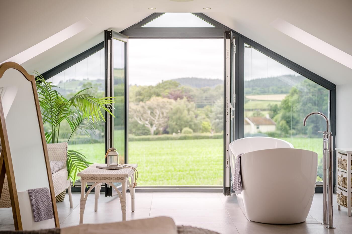 A modern room with a skylight, a bathtub, a table with a lantern, and a view of a green landscape.