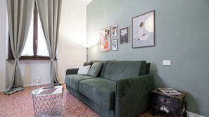 Ein Wohnzimmer mit einem grünen Sofa, einem kleinen Beistelltisch, einer Stehlampe und Kunstwerken an der Wand.
