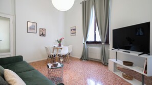 Ein Wohnzimmer mit einem grünen Sofa, einem Esstisch mit Stühlen, einem Fernseher auf einem Schrank und einer Heizung.