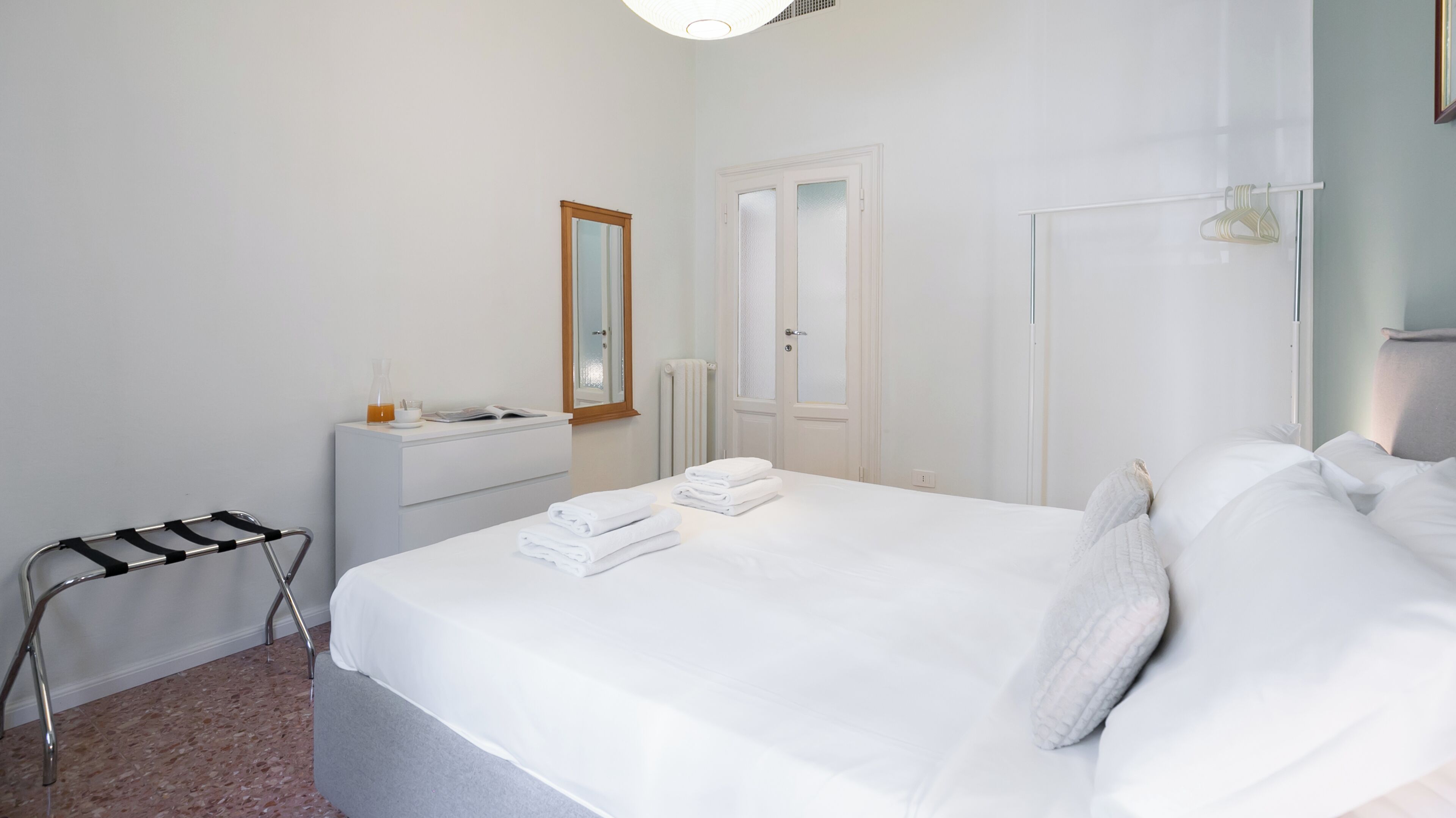 A hotel room with a bed, a bedside table, a radiator, a mirror, and a hanging light fixture.
